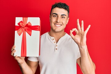 Hispanic young man holding gift doing ok sign with fingers, smiling friendly gesturing excellent symbol