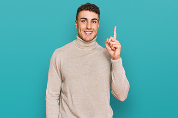 Fototapeta premium Hispanic young man wearing casual turtleneck sweater pointing finger up with successful idea. exited and happy. number one.