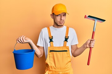 Hispanic young man wearing glass cleaner uniform and squeegee clueless and confused expression....