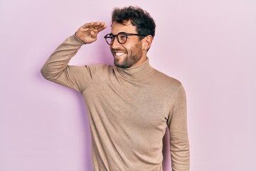 Handsome man with beard wearing turtleneck sweater and glasses very happy and smiling looking far away with hand over head. searching concept.