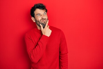 Handsome man with beard wearing casual red sweater thinking worried about a question, concerned and nervous with hand on chin