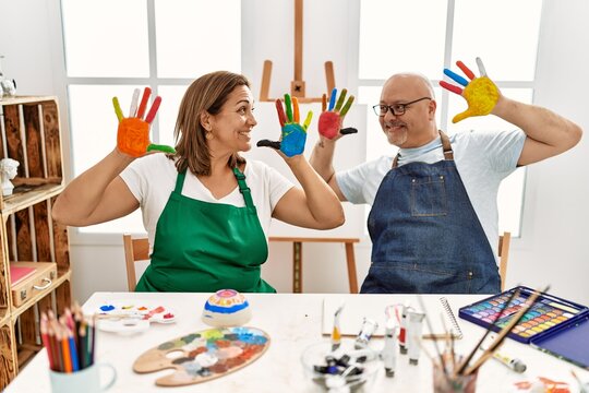 Middle age hispanic painter couple smiling happy showing painted palm hands at art studio.
