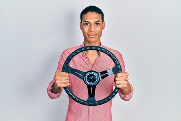 Young african american guy holding steering wheel relaxed with serious expression on face. simple and natural looking at the camera.