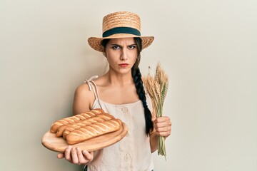 Young hispanic woman holding homemade bread and spike wheat skeptic and nervous, frowning upset...