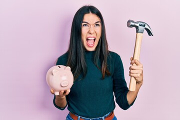 Young brunette woman holding piggy bank and hammer angry and mad screaming frustrated and furious, shouting with anger looking up.