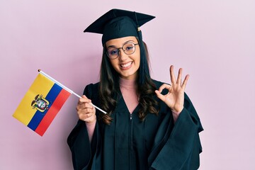 Young hispanic woman wearing graduation uniform holding ecuador flag doing ok sign with fingers, smiling friendly gesturing excellent symbol