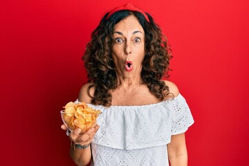 Middle age hispanic woman holding potato chip scared and amazed with open mouth for surprise, disbelief face