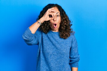 Middle age hispanic woman wearing casual clothes doing ok gesture shocked with surprised face, eye looking through fingers. unbelieving expression.