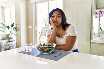 Young hispanic woman eating healthy salad at home thinking looking tired and bored with depression...