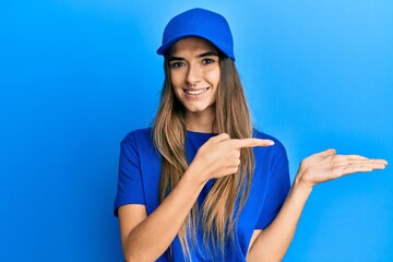 Fototapeta premium Young hispanic woman wearing delivery uniform and cap amazed and smiling to the camera while presenting with hand and pointing with finger.