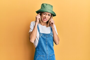 Young caucasian blonde woman wearing denim jumpsuit and hat with 90s style covering ears with...