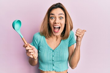 Young caucasian blonde woman holding silicone soup spoon pointing thumb up to the side smiling...