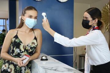 Receptionist and guest wearing face mask at front desk while having conversation in office or...