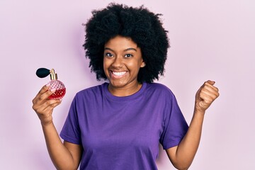 Young african american woman holding perfume screaming proud, celebrating victory and success very...