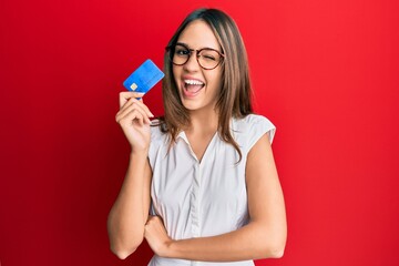 Young brunette woman holding credit card winking looking at the camera with sexy expression, cheerful and happy face. - obrazy, fototapety, plakaty