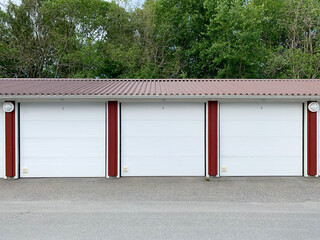Three garage doors, parking lots, with space for cars. White gates, paved road and trees.