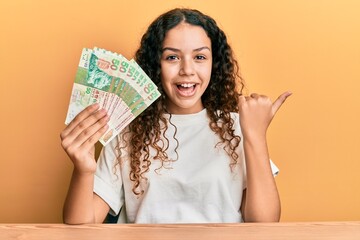 Teenager hispanic girl holding 50 hong kong dollars banknotes pointing thumb up to the side smiling happy with open mouth