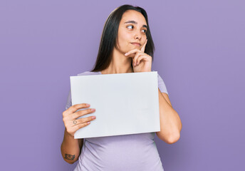 Young hispanic woman holding blank empty banner serious face thinking about question with hand on chin, thoughtful about confusing idea