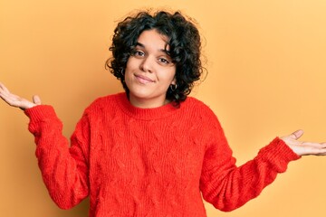 Young hispanic woman with curly hair wearing casual winter sweater clueless and confused expression with arms and hands raised. doubt concept.