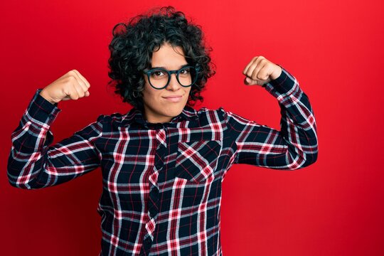 Young hispanic woman with curly hair wearing casual clothes and glasses showing arms muscles smiling proud. fitness concept.