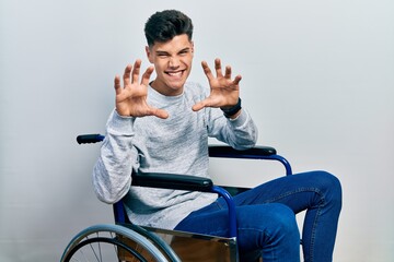 Young hispanic man sitting on wheelchair smiling funny doing claw gesture as cat, aggressive and...