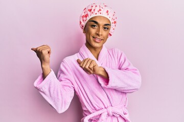 Hispanic man wearing make up wearing shower towel cap and bathrobe pointing to the back behind with hand and thumbs up, smiling confident