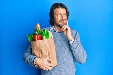 Middle age handsome man holding paper bag with groceries serious face thinking about question with...