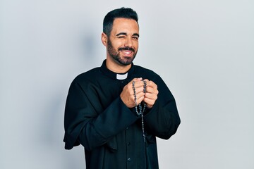 Handsome hispanic priest man with beard praying holding catholic rosary winking looking at the...