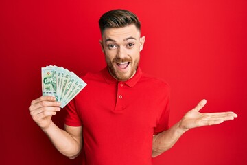 Young redhead man holding 20 thai baht banknotes celebrating achievement with happy smile and winner expression with raised hand