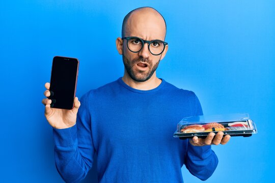 Young hispanic man holding take away food showing smartphone screen in shock face, looking skeptical and sarcastic, surprised with open mouth