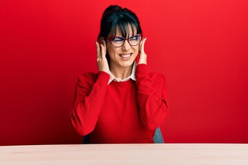Young brunette woman with bangs wearing glasses sitting on the table covering ears with fingers with annoyed expression for the noise of loud music. deaf concept.