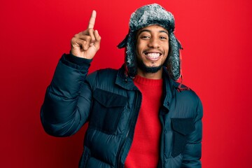 Young african american man with beard wearing winter hat and coat pointing finger up with successful idea. exited and happy. number one. - obrazy, fototapety, plakaty