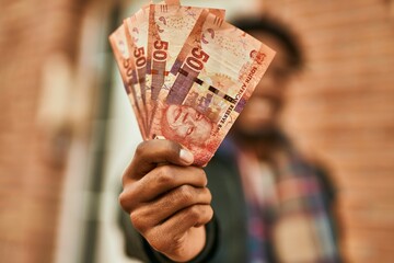 Young african american man smiling happy  holding south africa rands banknotes at the city