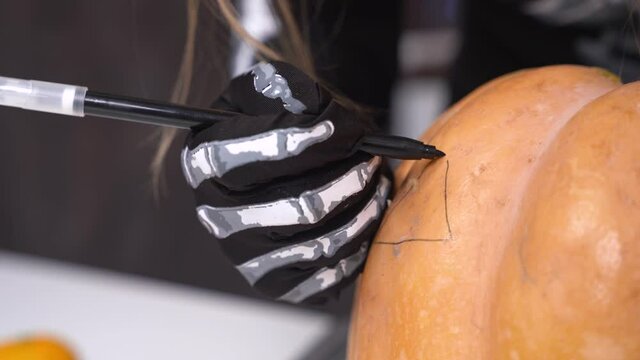 Close up human hands wearing black gloves with skeleton drawing the mockup on big pumpking for cutting, with marker. Halloween concept.