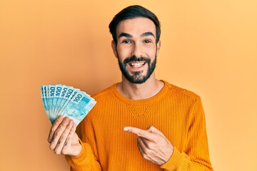 Young hispanic man holding 100 brazilian real banknotes smiling happy pointing with hand and finger