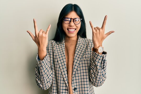 Young chinese woman wearing business style and glasses shouting with crazy expression doing rock symbol with hands up. music star. heavy music concept.