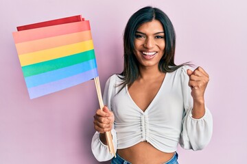 Young latin transsexual transgender woman holding rainbow lgbt flag screaming proud, celebrating victory and success very excited with raised arm
