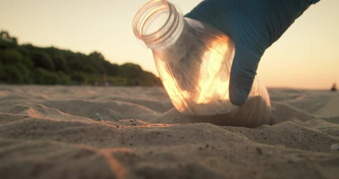 Close-up of the hand of a volunteer in a rubber glove, picking up a plastic bottle from a sandy beach. The concept of plastic pollution and environmental problems. Voluntary cleaning of nature from pl