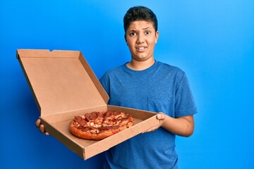 Teenager hispanic boy eating tasty pepperoni pizza clueless and confused expression. doubt concept.