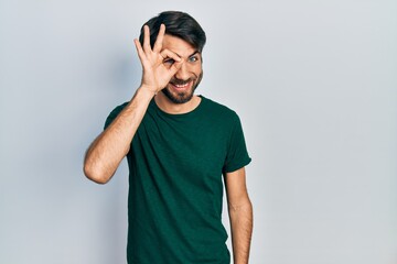 Young hispanic man wearing casual white tshirt doing ok gesture with hand smiling, eye looking through fingers with happy face.