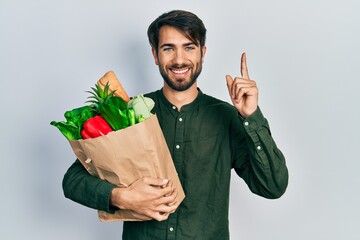 Young hispanic man holding paper bag with bread and groceries surprised with an idea or question...