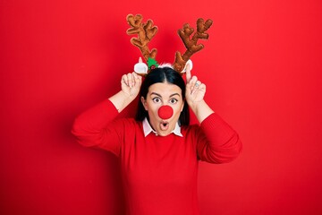 Young hispanic woman wearing deer christmas hat and red nose doing funny gesture with finger over...