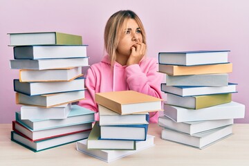 Young caucasian woman sitting on the table with books looking stressed and nervous with hands on mouth biting nails. anxiety problem.