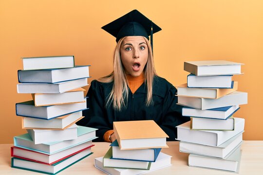 Young caucasian woman wearing graduation ceremony robe sitting on the table afraid and shocked with surprise and amazed expression, fear and excited face.