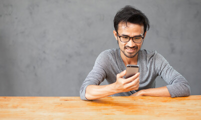 Fototapeta premium happy young man smiling and using mobile phone with wooden table and concrete wall background and copy space