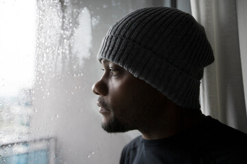 Closeup portrait of a sad African American man looking out through glass window on a wet cold rainy...