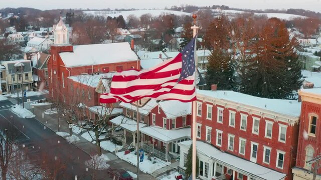 Small town America in winter. USA flag waves with snow flakes. Home, business, house apartment view. Aerial drone shot. Christmas holiday season.