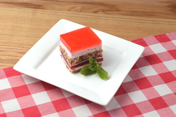 A delicious looking dessert cake served on a white plate. Layered sponge cake with jello layers and mint leaf on top.