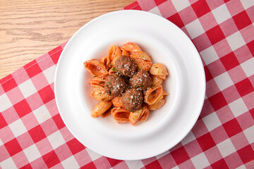 Penne pasta with meatballs in tomato sauce in a white bowl. Top view. On rustic wooden table with red plaid tablecloth.