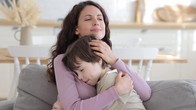 Happy mother kiss cute boy hugging mom in temple with closed eyes expressing tenderness, love and care. Family unity. Caring mom enjoy embracing son sitting on sofa at home on weekend or after work